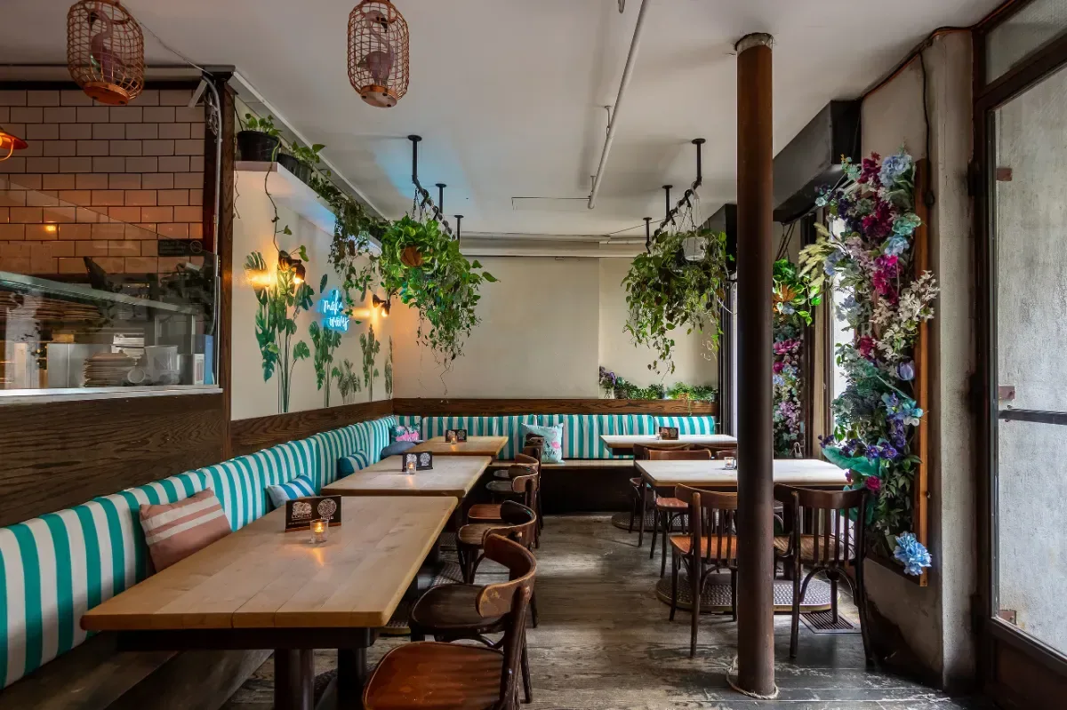 Loreley Beer Garden's Garden Room in daylight, with teal and white striped banquettes, wooden tables, hanging plants and a wall of flowers