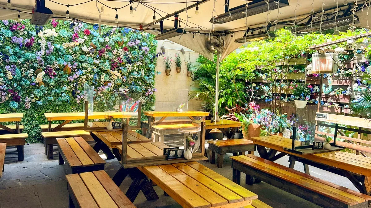 Wooden picnic tables under a canopy and string lights in front of a flower wall at Loreley Beer Garden
