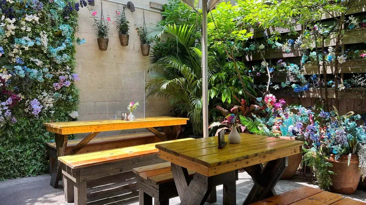 Picnic tables in the garden at Loreley Beer Garden under green plants, hanging baskets and a wall of blue and white flowers