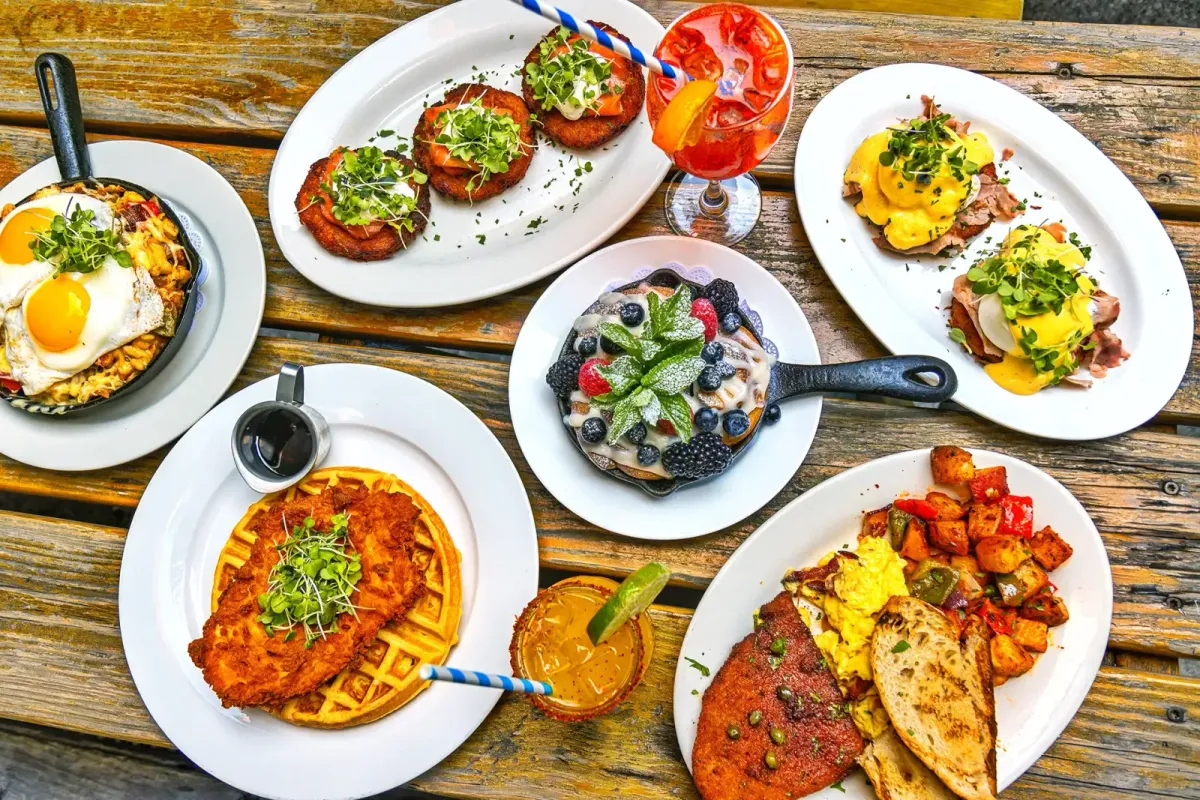 A brunch table seen from above, with eggs in a skillet, a chicken and waffle, eggs Benedict and cocktails on a wooden table