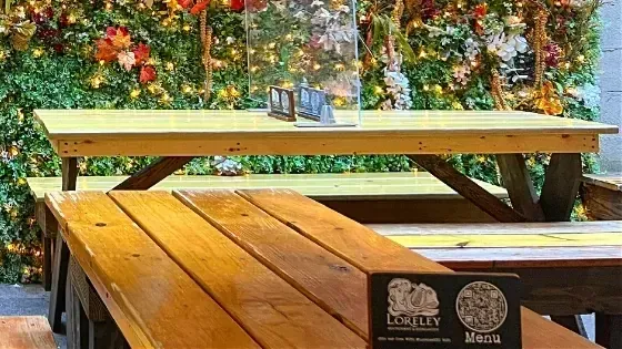 A small Loreley menu card with a code standing at the end of a wooden picnic table, with a flower wall behind